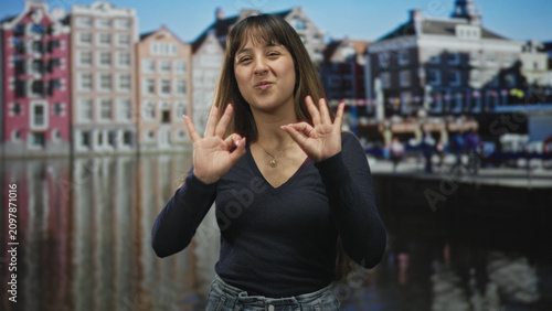 Фотография Young hispanic woman shows two ok hand gestures with visible hands and gold necklace, smiling before a canal building backdrop in studio; joyful confidence