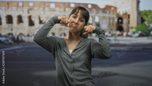 Young woman pinches her cheek and pulls down her mouth with fingers in front of an ancient building on a busy street; self mockery frustration.