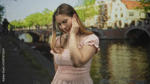 Slika na platnu Woman touching ear with hand on canal street by arched bridge, wearing pink dress and looking down; discomfort concern