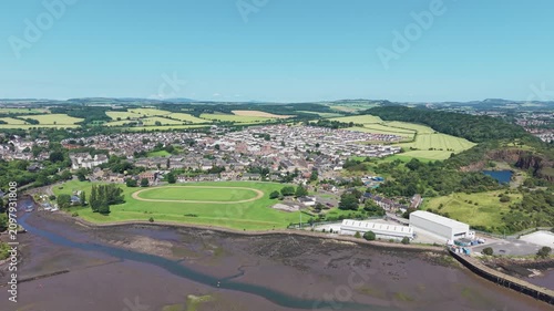 Aerial view of the coastal village of Inverkeithing in Fife, Scotland, flying towards the residential area, green sports field, and muddy bay during a bright summer day