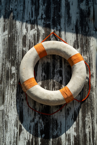 A white and orange circular life preserver hangs on a weathered wooden wall. It is designed as an emergency flotation device for safety in the ocean or pool.