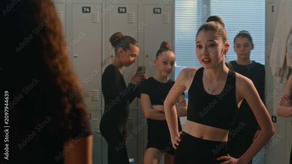 Fototapeta premium In Sports Locker Room, Young Female Mock, Intimidate Curly Haired Teammate While One Girl Records Incident on Smartphone. Bullying, Exclusion, Toxic Behavior in Youth Sports and Teenage Peer Culture.