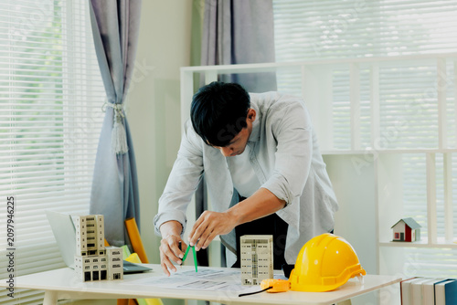 Contented Asian corporate engineer verifying structural project blueprints at bright modern desk.
