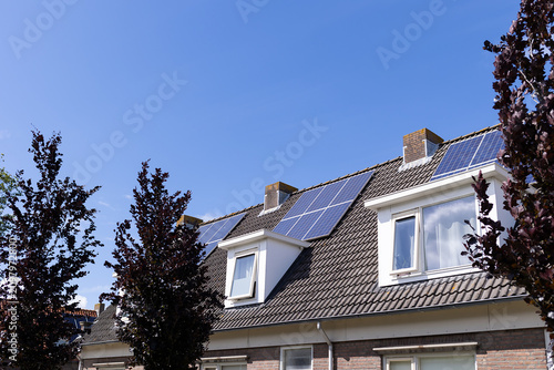 Close view of tiled rooftop fitted with multiple solar panels beside brick chimneys and white dormer windows, framed by dark trees under bright blue sky, showing sustainable energy use