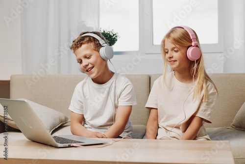 Two smiling children with headphones watch a laptop while sitting on a beige sofa in a bright living room. Concept of online learning  digital entertainment  and childhood technology.