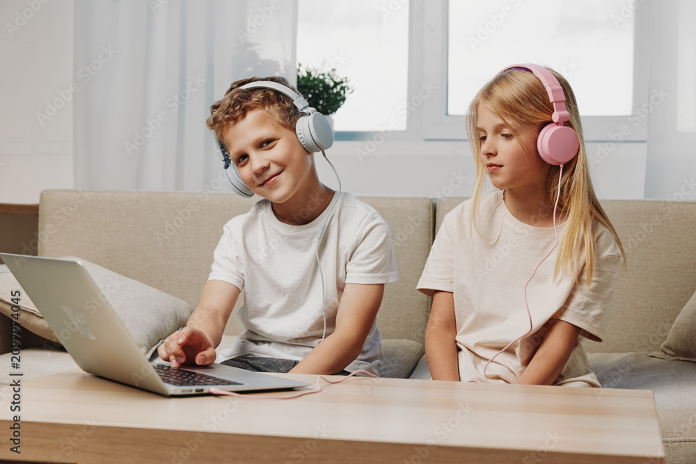 Obraz premium Two children with headphones use laptop at home sitting on couch. Concept of remote learning, entertainment, and digital technology for kids in casual room setting.