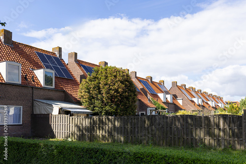 Sunlit row of brick homes with steep terracotta roofs, rooftop solar arrays, dormer windows, brick chimneys, green trees and wooden fence, creating quiet residential streetscape, with solar, roof