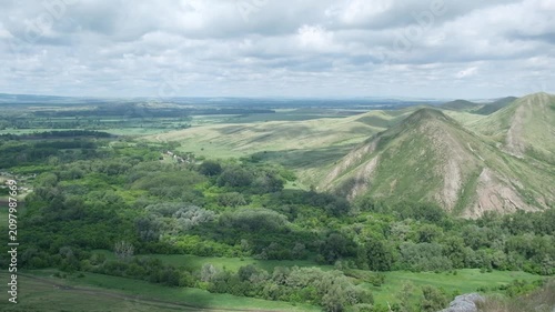 Expansive panoramic landscape with rolling green hills, steep grassy ridges, dense woodland, layered valleys, rocky foreground, and broad cloudy sky under soft daylight