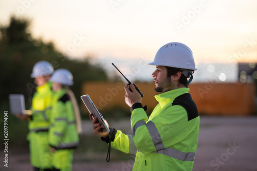 Fotografie Construction professionals in safety gear collaborating at sunset using tablets