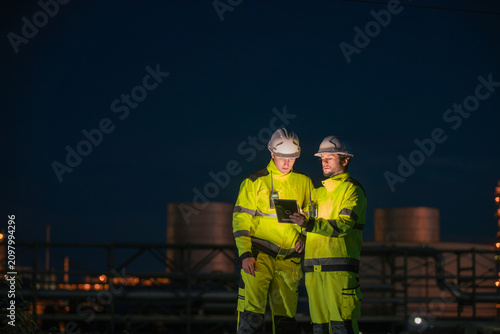 Engineers reviewing data on a tablet at a construction site during twilight with industrial structures in the background
