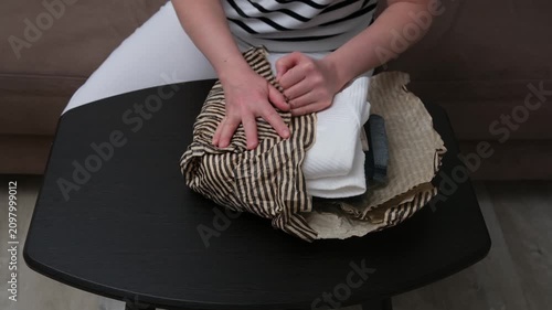 Person seated on brown sofa folding and packing striped clothing on small black table in calm home interior with soft natural light, neutral cushions, beige floor, and centered domestic composition