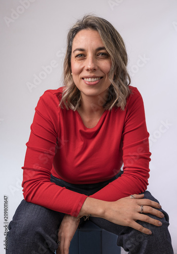 Friendly woman sitting casually and smiling at the camera against a neutral background, representing confidence, authenticity, wellbeing and approachability.