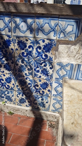 Decorative blue and white ceramic tiles featuring intricate floral patterns on a wall with a visible stone edge and sunlight casting shadows