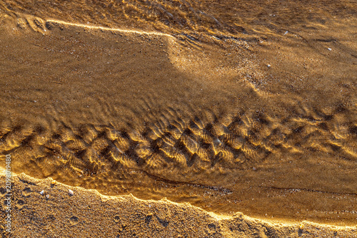 Golden sunset light reflecting on wet sand pattern made by backwash water stream. Beautiful mediterranean beach shore texture with warm sun reflection on ripple in summer