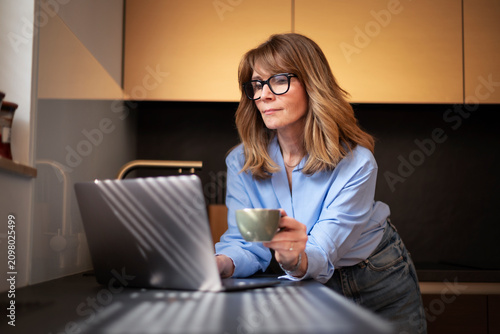 Blond haired smiling woman with laptop standing in a modern kitchen and working from home