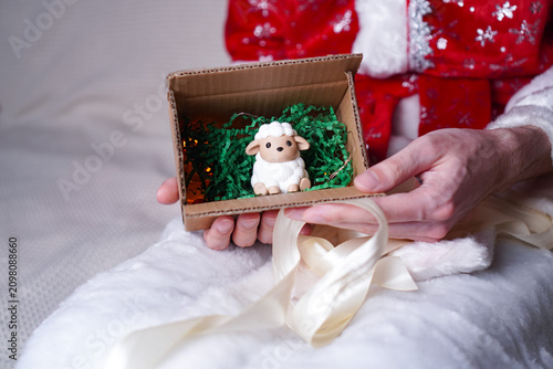 a man in a Santa Claus costume holding a box with an unpacked gift in his hands, and in another a vintage figure of a white sheep symbol of 2027 on the background of a white sofa as a New Year card 