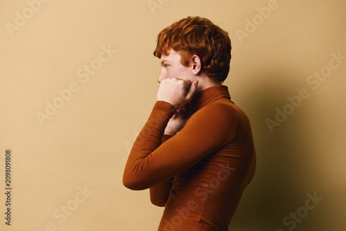 Tableau sur toile Profile of redhead woman with short curly hair wearing brown turtleneck, standing with closed fists near face against beige background indoor