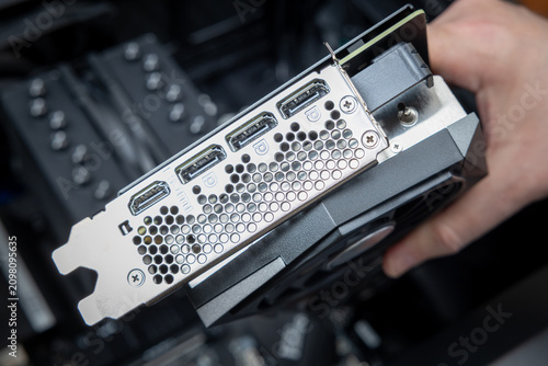 A technician's hand holds a powerful graphics card above a motherboard during gaming PC assembly. The silver rear bracket has three DisplayPort ports and one HDMI port