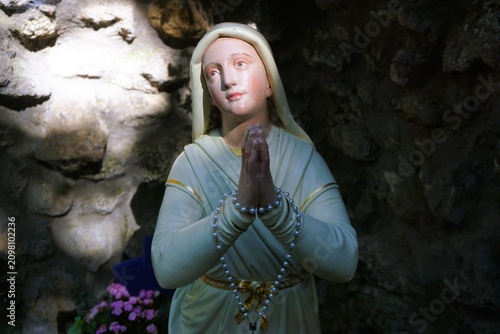 Fotografía Statue von betender Heiliger Bernadette vor Fels in Grotte bei Sonne am Abend im