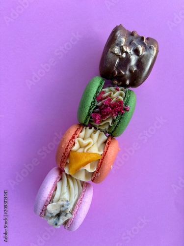Flat lay of four unique gourmet macarons arranged in a vertical line on a vibrant purple background. Features artisanal flavors including a chocolate-glazed gold shell, a green macaron with raspberry 
