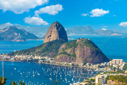 Stunning aerial view of Sugarloaf Mountain and Guanabara Bay in Rio de Janeiro, Brazil, featuring anchored boats and a vibrant blue sky on a sunny day.