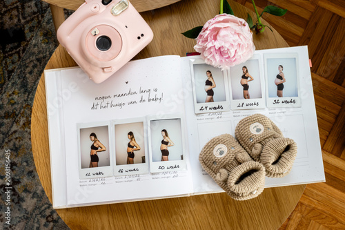 Фотография A pregnancy journal filled with instant photos of the growing belly lies open with knitted baby booties a pink peony and an instant camera a tender maternity flat lay