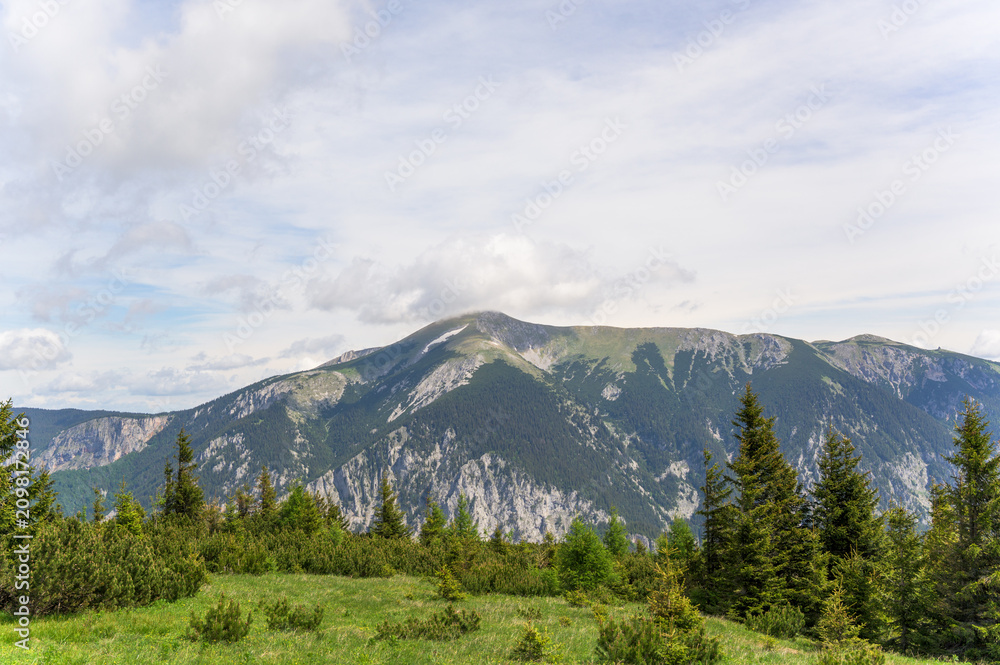Obraz premium A breathtaking view of Rax Mountain in Austria featuring rolling green hills and rugged peaks under a vibrant cloudy sky with soft sunlight illuminating the lush greenery