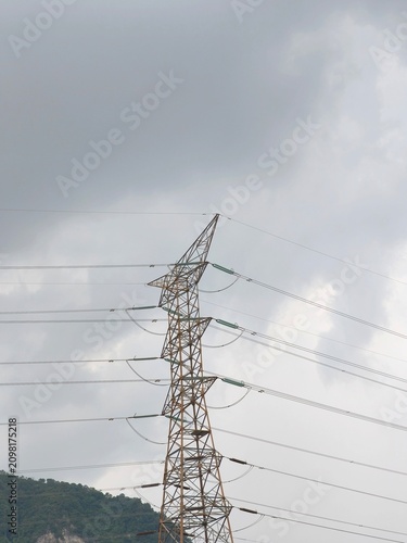 Detailed view of electricity transmission tower constructed from steel lattice beams supporting insulators and high-voltage lines against cloudy sky and hillside backdrop