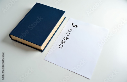 Blue hardbound book sits next to white paper sheet on desk. Printed document contains tax regulation checklist with checked boxes. Office pro examines financial statement for legal compliance.