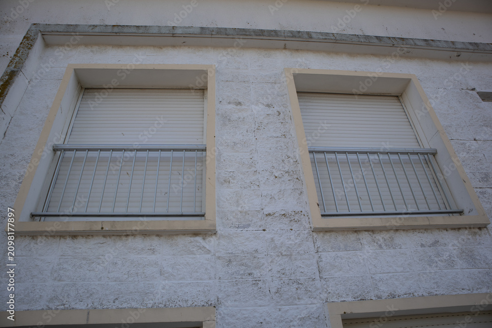 Fototapeta premium Two closed windows with roller shutters and safety railings on weathered facade