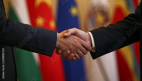 Two politicians in dark suits shaking hands firmly with flags in the background