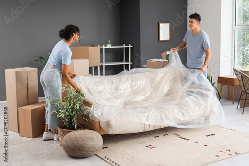 Young couple covering sofa with film in room on moving day