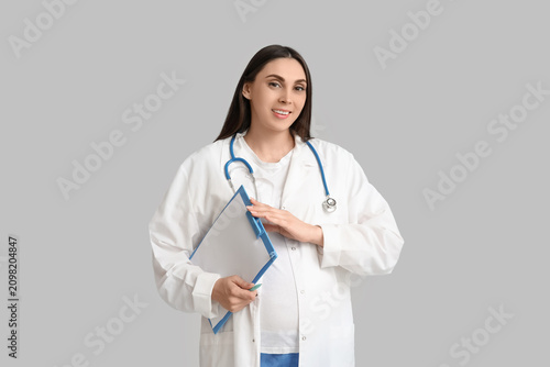 Beautiful young pregnant doctor with clipboard and stethoscope on grey backgr...