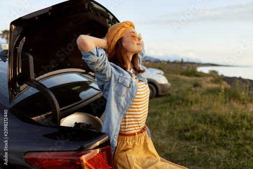 Obraz na plátne Young woman in striped tee and denim jacket leans against a car trunk, eyes closed, basking in a warm breeze during golden hour, carefree and relaxed