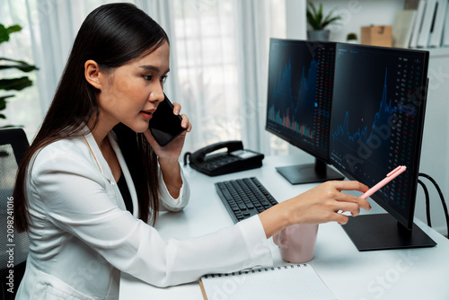 Foto Young beautiful Asian businesswoman calling to broker, pointing with pen in increased exchange stock market on pc to invest high profit at modern office, investing high profit in wealth