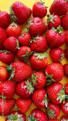 Vertical macro shot of a background completely filled with fresh, juicy red strawberries with green leaves over a yellow surface. Close-up pattern of organic summer berries. Vibrant food flat lay temp