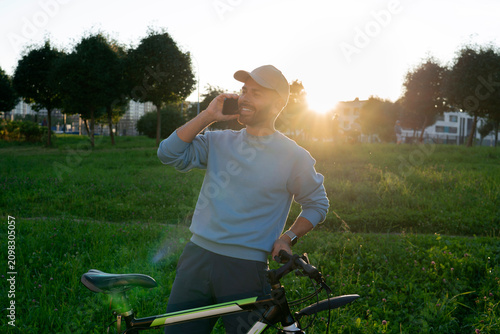 Cheerful guy enjoys a peaceful summer morning in a city park while talking on a smartphone and standing next to his bicycle under the warm sunrise light
