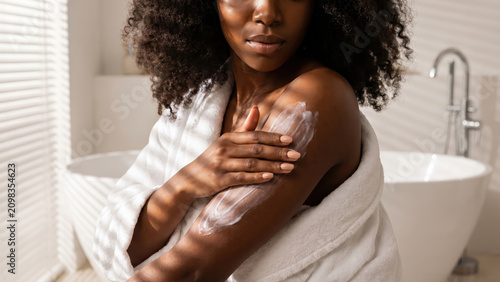 Black woman applying moisturizing body lotion for healthy skin in a bright bathroom.