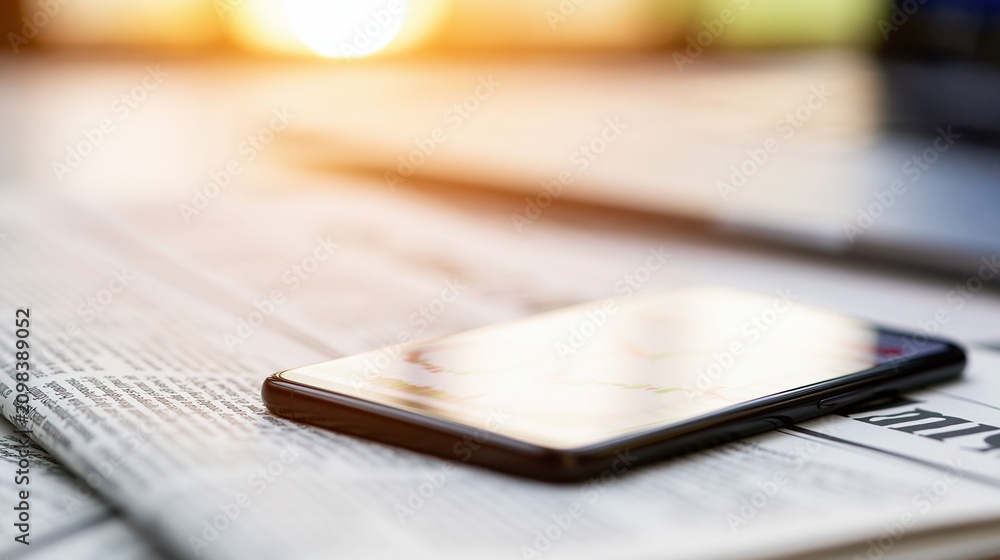 Naklejka premium Modern Smartphone Resting on Newspaper with Blurred Sunlight Glare - Business News Update