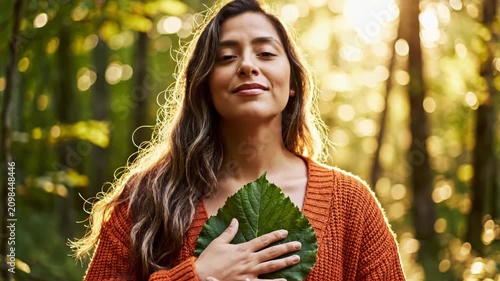 Woman in orange cardigan holding green leaf to her heart with eyes closed in a sunlit forest at golden hour, focus on nature connection and mindfulness in the autumn woods.