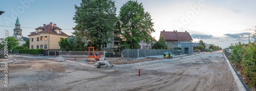 A wide panoramic view captures a major road reconstruction project in Siska, Ljubljana. The gravel-covered street is flanked by residential houses, construction machinery like rollers 