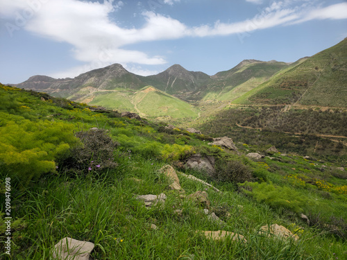 Lush spring mountain landscape with green hills, orchards, rocky terrain, wild vegetation, and dramatic peaks beneath a partly cloudy blue sky.
📍 Hawraman Takht, Kurdistan, Iran 