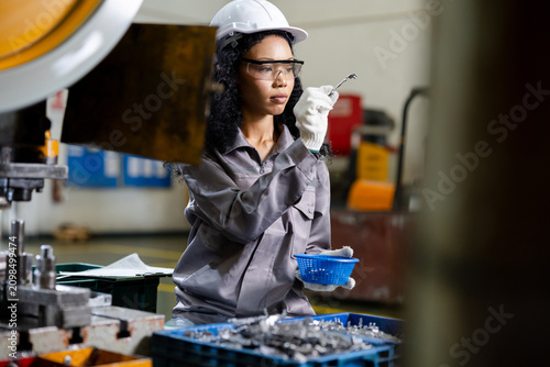 Factory technician wearing safety gear inspects a small metal part beside industrial machinery, holding a bin of fasteners in a busy manufacturing workspace.