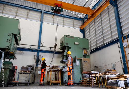 Industrial workshop with two engineers in safety gear inspecting heavy machinery beneath an overhead crane inside a metal fabrication plant.