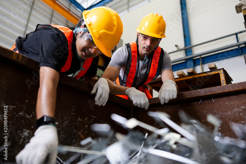 Two factory workers in hard hats and safety vests sort sharp metal scrap in a large bin at an industrial recycling facility, with motion blur showing active processing.