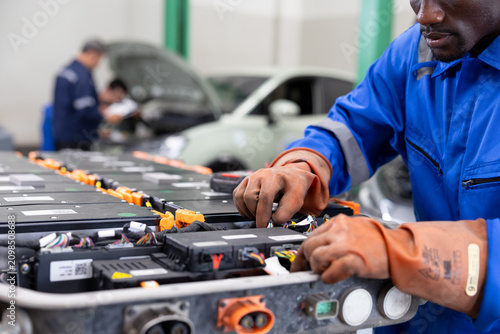 Konstfotografi Technician wearing protective gloves services an electric vehicle battery pack in an auto workshop, inspecting modules and connections with tools beside a car