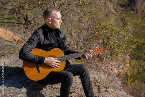 Blonde man playing string guitar outdoors in autumn forest. Concept of sound therapy, mental health and wellness rituals. Calmness tranquility audio-sensory practices. Aura farming energy holistic