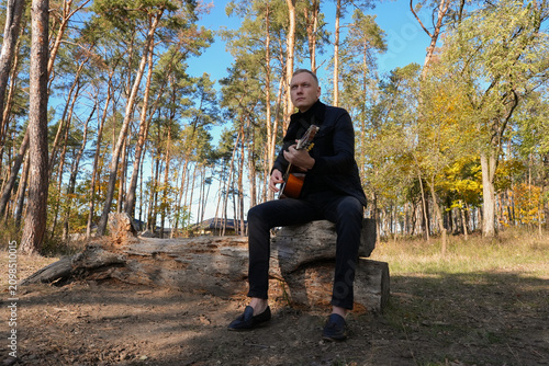 Beautiful blonde man playing acoustic guitar and singing relaxing enjoying outdoors on sunny autumn day. Artist learning and practicing playing musical instrument. Fall leaves autumnal weather 