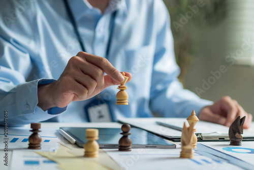 Close-up of businessman holding chess piece planning strategy at desk with digital tablet and charts.