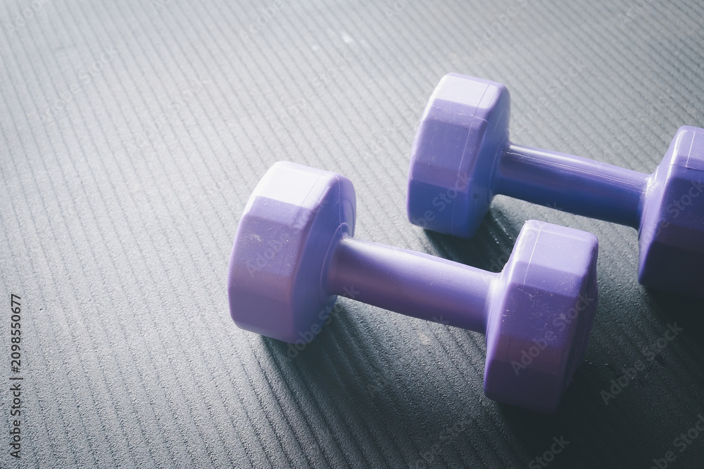Obraz premium Close up of two purple dumbbells resting on a black textured gym mat with natural light. Home workout, strength training, weight loss, and active fitness lifestyle concept with copy space.